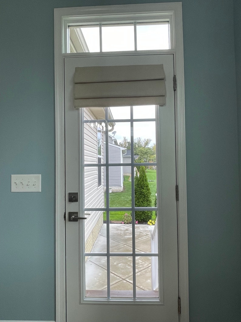 May include: A white door with a grid of glass panes, offering a view of a garden with green grass and a small tree. A beige Roman blind is mounted above the glass. The door is set in a light blue wall.