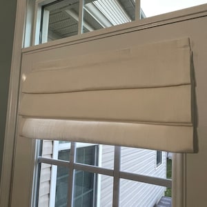 Puede incluir: Una cortina romana de color crema est&aacute; instalada en una ventana con un patr&oacute;n de rejilla. La cortina est&aacute; hecha de una tela texturizada y est&aacute; parcialmente bajada, revelando la ventana y la vista exterior. El marco de la ventana es blanco.
