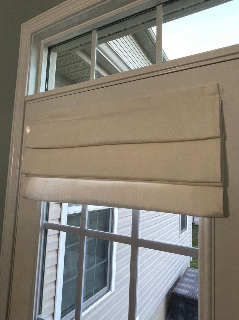 Puede incluir: Una cortina romana de color crema est&aacute; instalada en una ventana con un patr&oacute;n de rejilla. La cortina est&aacute; hecha de una tela texturizada y est&aacute; parcialmente bajada, revelando la ventana y la vista exterior. El marco de la ventana es blanco.
