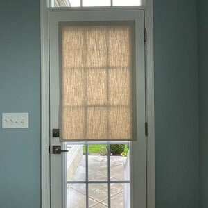 May include: A white door with a window and a beige linen shade. The door has a small window at the top and a larger window with a grid pattern. The door is set in a white frame against a light blue wall.