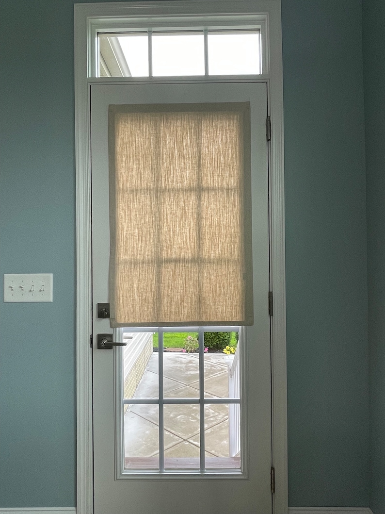 May include: A white door with a window and a beige linen shade. The door has a small window at the top and a larger window with a grid pattern. The door is set in a white frame against a light blue wall.