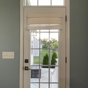 Puede incluir: Una puerta blanca con una rejilla de cristales, coronada por una ventana rectangular y una cortina romana color crema. La puerta tiene un tirador y bisagras plateadas. La vista a trav&eacute;s del cristal muestra un jard&iacute;n con hierba verde y &aacute;rboles.