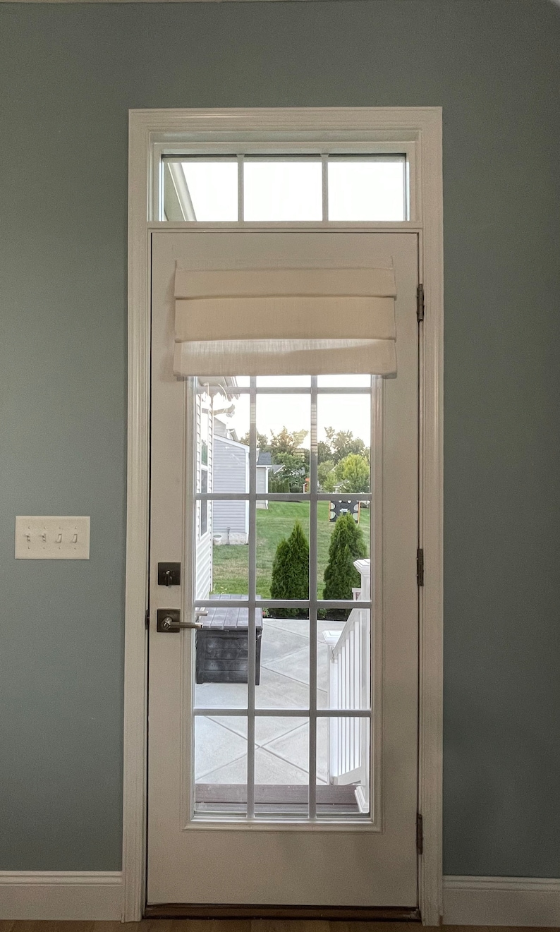 Puede incluir: Una puerta blanca con una rejilla de cristales, coronada por una ventana rectangular y una cortina romana color crema. La puerta tiene un tirador y bisagras plateadas. La vista a trav&eacute;s del cristal muestra un jard&iacute;n con hierba verde y &aacute;rboles.