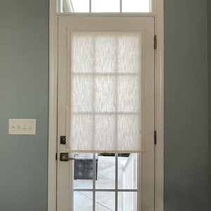 Puede incluir: Una puerta blanca con un dise&ntilde;o de panel de ventana rectangular y una persiana de tela blanca. La puerta est&aacute; colocada en un marco blanco contra una pared azul claro. Una peque&ntilde;a ventana est&aacute; encima de la puerta.