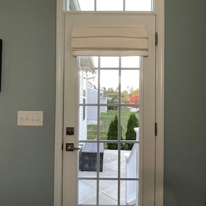 Puede incluir: Una puerta francesa blanca con un patr&oacute;n de rejilla, coronada por una ventana rectangular. Una cortina romana color crema est&aacute; parcialmente bajada. La puerta se abre a un patio con un banco y una vista de un c&eacute;sped verde y &aacute;rboles.