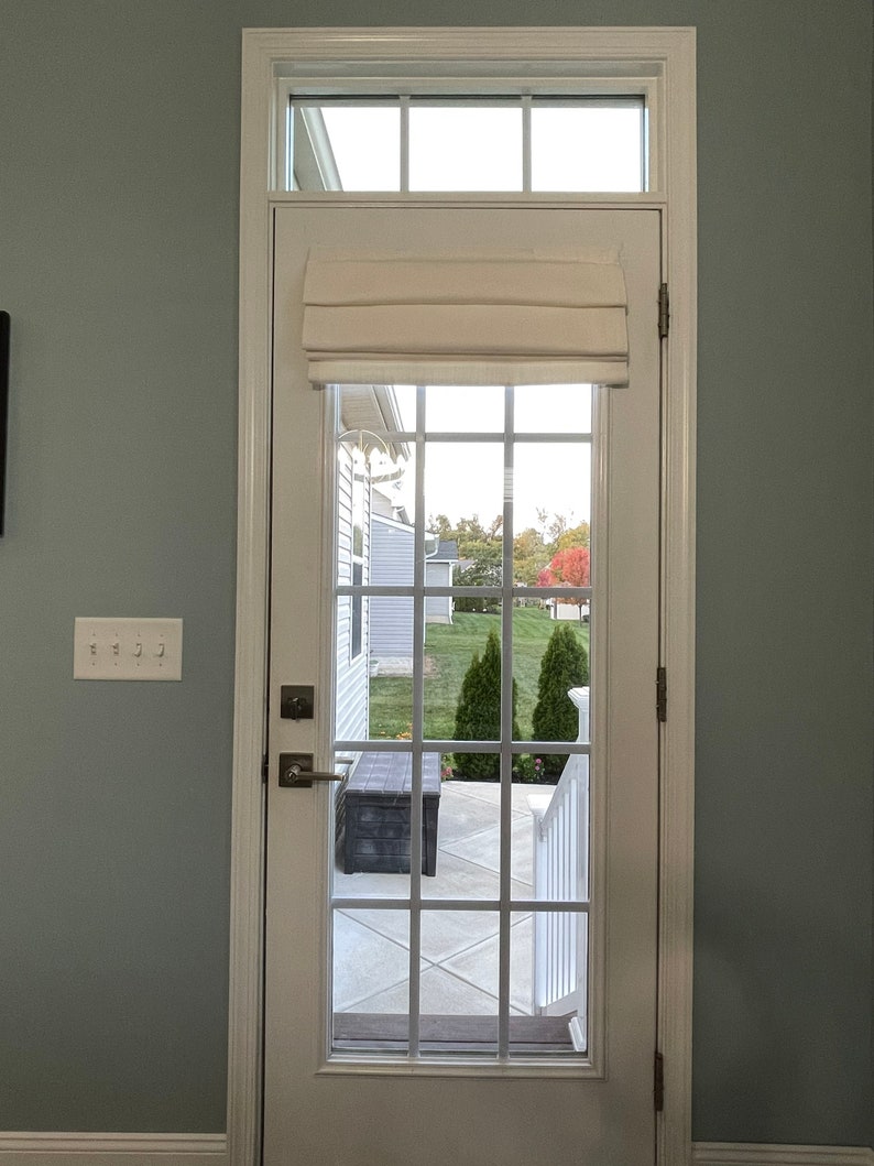 Puede incluir: Una puerta francesa blanca con un patr&oacute;n de rejilla, coronada por una ventana rectangular. Una cortina romana color crema est&aacute; parcialmente bajada. La puerta se abre a un patio con un banco y una vista de un c&eacute;sped verde y &aacute;rboles.