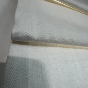 Puede incluir: Primer plano de una tela blanca texturizada, posiblemente lino o lona, ​​doblada en capas. La tela tiene un patr&oacute;n sutil y parece ser parte de un tratamiento de ventana, como una persiana romana. Los pliegues crean sombras y profundidad.