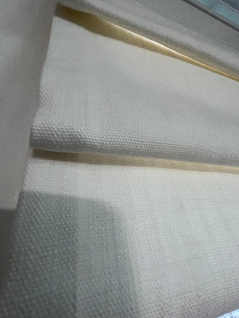Puede incluir: Primer plano de una tela blanca texturizada, posiblemente lino o lona, ​​doblada en capas. La tela tiene un patr&oacute;n sutil y parece ser parte de un tratamiento de ventana, como una persiana romana. Los pliegues crean sombras y profundidad.