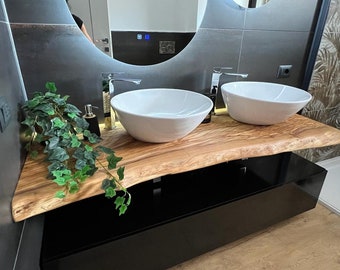 May include: A bathroom vanity with a wooden countertop and two white porcelain sinks. The countertop is made of a natural wood slab with a live edge. The vanity is black and has a drawer. A green plant is hanging over the countertop.