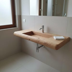 Könnte beinhalten: Ein modernes, an der Wand befestigtes Badezimmerwaschbecken aus hellem Holz. Das Waschbecken hat ein rechteckiges Becken und eine abgerundete Kante. Ein verchromter Wasserhahn und ein Abflussrohr sind sichtbar. Ein kleines, weißes Waschtuch liegt am Rand.