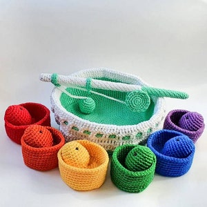 Puede incluir: Un conjunto de juguetes de crochet con una cesta verde y blanca, una caña de pescar y seis cestas de colores. Las cestas son roja, naranja, amarilla, verde, azul y morada.