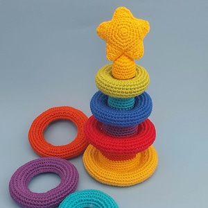 Op de afbeelding: Een regenboogkleurig gehaakt stapelspel met een gele ster bovenop. Het speelgoed is gemaakt van zacht garen en is perfect voor baby's en peuters.