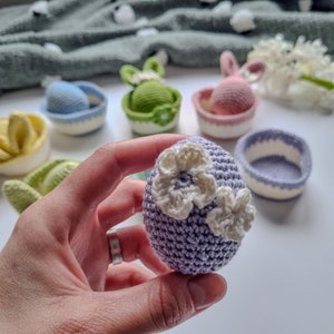 May include: A purple crocheted Easter egg with white crocheted flowers. The egg is being held in a hand.