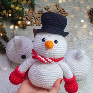 Op de afbeelding: Een witte gehaakte sneeuwpop met een zwarte hoge hoed, een rood-wit gestreepte sjaal en rode wanten. De sneeuwpop zit op een wit oppervlak.