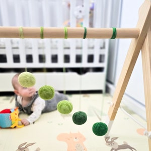 Könnte beinhalten: Ein Baby-Spielbogen aus Holz mit fünf grünen gehäkelten Bällen, die an der oberen Stange hängen. Die Bälle sind in verschiedenen Grüntönen gehalten, von hell bis dunkel. Der Spielbogen ist auf einer Spielmatte mit hellbraunem Hintergrund aufgebaut.