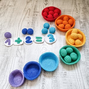 Peut inclure: Un ensemble de paniers en crochet colorés remplis de petites boules en crochet. Les paniers sont disposés en arc-en-ciel, avec les couleurs rouge, orange, jaune, vert, bleu et violet. Les boules sont également en crochet de différentes couleurs, notamment blanc, vert et bleu. Les paniers sont étiquetés avec des chiffres et des symboles mathématiques, ce qui en fait un jouet amusant et éducatif pour les enfants.