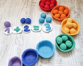 May include: A set of colourful crocheted baskets filled with small crocheted balls. The baskets are arranged in a rainbow pattern, with the colours red, orange, yellow, green, blue, and purple. The balls are also crocheted in different colours, including white, green, and blue. The baskets are labelled with numbers and mathematical symbols, making this a fun and educational toy for children.