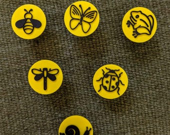 May include: Six circular yellow buttons, each featuring a distinct black embossed insect or animal graphic. The designs include a bee, a butterfly, a frog, a dragonfly, a ladybug, and a snail, all arranged in a scattered pattern on a textured surface.