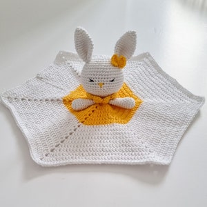 May include: A white crocheted baby blanket with a yellow hexagon in the center. A white crocheted bunny with a yellow bow sits in the center of the hexagon.
