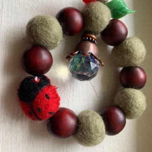 Könnte beinhalten: Ein roter und schwarzer Marienkäfer-Anhänger hängt an einem Kranz aus braunen Holzperlen und grünen Filzkugeln. Ein Kristallprisma und eine Metallglocke hängen an dem Kranz.