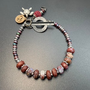 Puede incluir: Un brazalete de piedra de ágata roja y blanca con cuentas y abalorios plateados. Los abalorios incluyen un símbolo yin yang, una tortuga y una pequeña roca de lava roja.