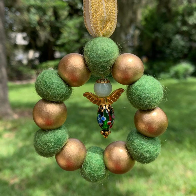 May include: A green and gold felt ornament with a small angel charm in the center. The angel is holding a green and white glass bead.