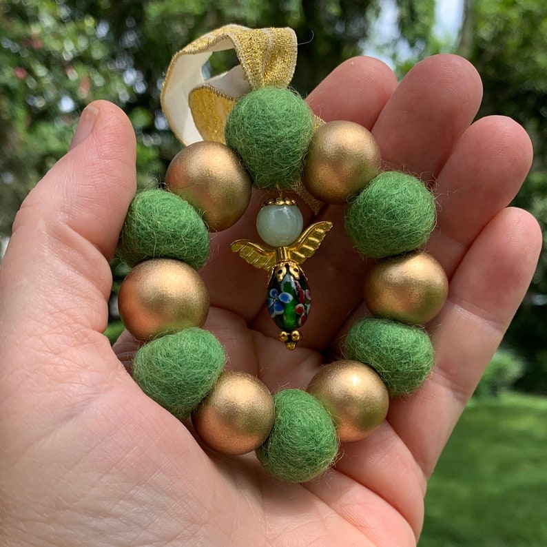 May include: A green and gold felt Christmas ornament with an angel charm. The ornament is made of felt balls and has a gold ribbon.