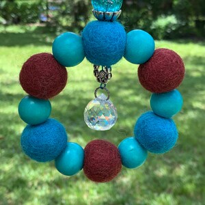 May include: A teal and maroon felt bead wreath with a crystal prism hanging from the center. The wreath is topped with a teal glass bead and a silver metal cap.