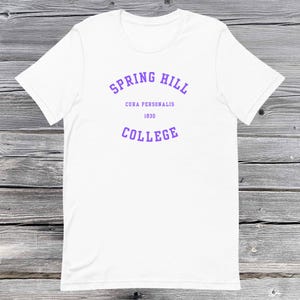 Op de afbeelding: Wit t-shirt met de paarse tekst "SPRING HILL COLLEGE", "CURA PERSONALIS" en het jaartal "1830". Het shirt is gemaakt van zacht materiaal en heeft korte mouwen.