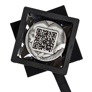 May include: A silver-colored, round, decorative object with a black and white QR code in the center. The object is surrounded by two hands, and the text "Audiobiography" is engraved below the QR code. The object is displayed in a black square box with a clear plastic cover.