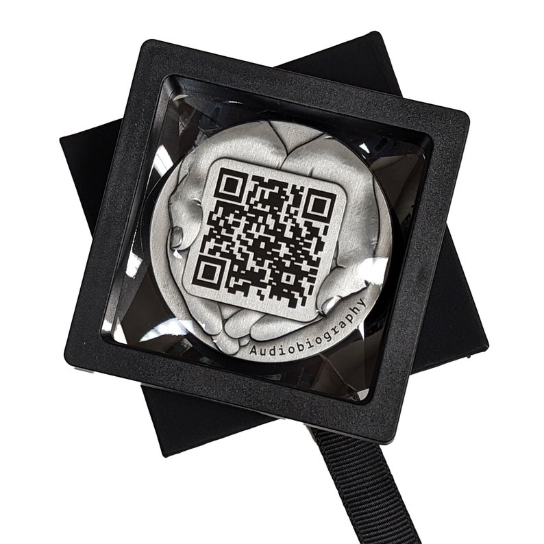 May include: A silver-colored, round, decorative object with a black and white QR code in the center. The object is surrounded by two hands, and the text "Audiobiography" is engraved below the QR code. The object is displayed in a black square box with a clear plastic cover.