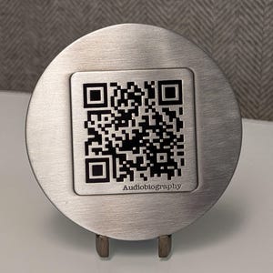 May include: A circular brushed silver metal plaque featuring a square black and white QR code centered on the surface. Below the QR code, the word Audiobiography is printed in a clear, sans-serif font. The plaque is supported by a small metal stand.