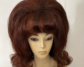 May include: A vintage auburn wig with a bouffant style, featuring a voluminous crown and curled ends. The wig is displayed on a mannequin head, showcasing its color and texture. The hair has a natural-looking sheen.