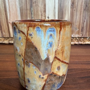 May include: A cylindrical ceramic mug featuring a variegated glaze with earthy tones of tan, dark brown, and ochre. The surface displays a crackled, mosaic-like pattern with subtle streaks of blue and white dripping down the exterior walls.