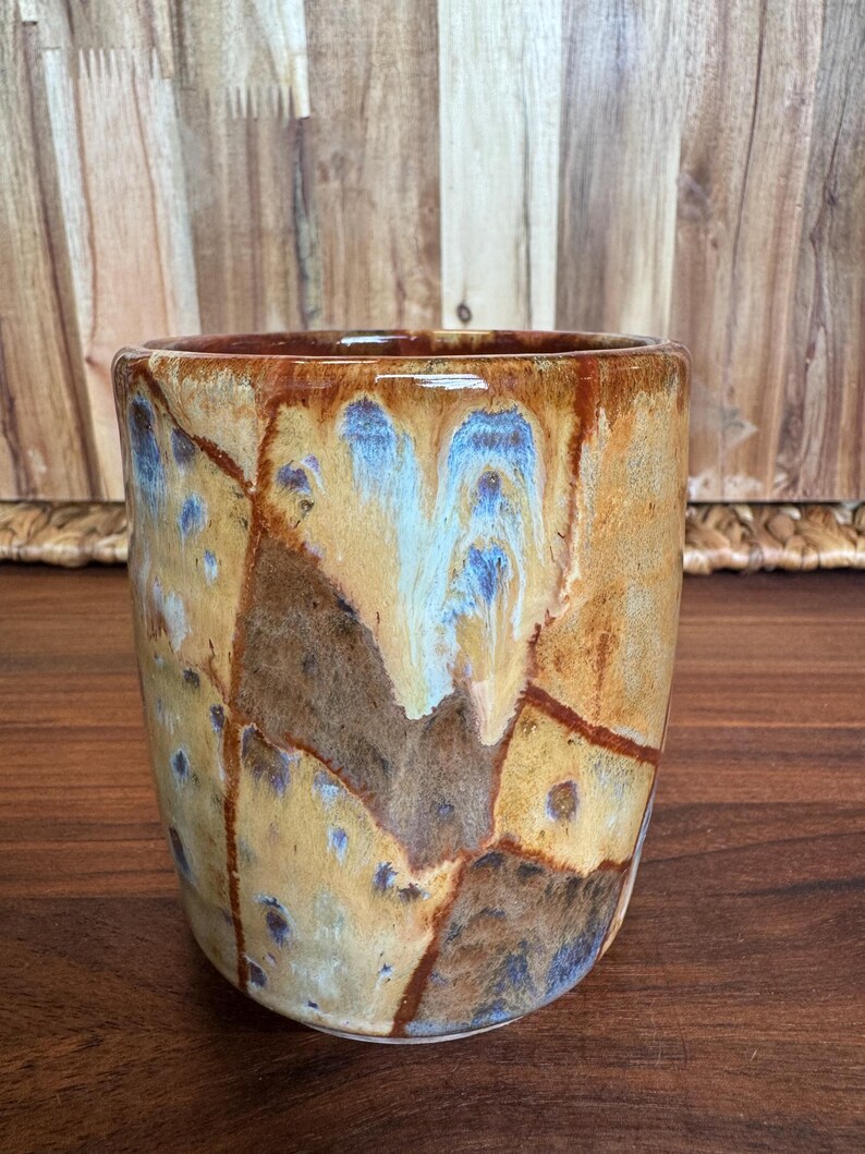 May include: A cylindrical ceramic mug featuring a variegated glaze with earthy tones of tan, dark brown, and ochre. The surface displays a crackled, mosaic-like pattern with subtle streaks of blue and white dripping down the exterior walls.