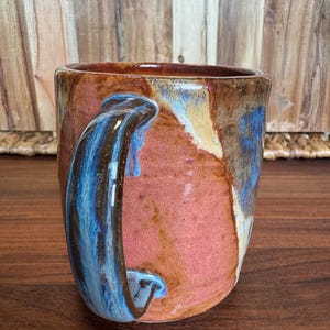 May include: A ceramic mug featuring a reactive glaze with blended tones of terracotta, deep blue, and cream. The mug has a sturdy, curved handle and a cylindrical body with a smooth, glossy finish that highlights the layered colour transitions.