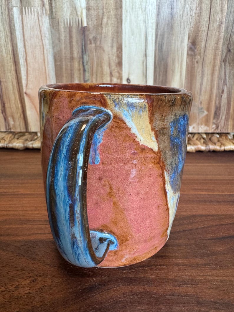 May include: A ceramic mug featuring a reactive glaze with blended tones of terracotta, deep blue, and cream. The mug has a sturdy, curved handle and a cylindrical body with a smooth, glossy finish that highlights the layered colour transitions.