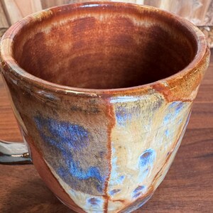 May include: A ceramic mug with a glossy finish featuring a blend of earthy brown, tan, and vibrant blue glaze. The interior is a solid reddish-brown tone, and the exterior displays a mottled, multi-coloured pattern with a smooth, reflective surface.