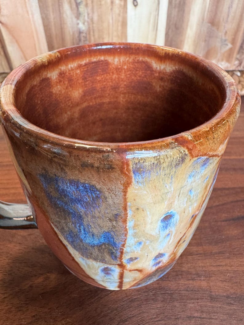 May include: A ceramic mug with a glossy finish featuring a blend of earthy brown, tan, and vibrant blue glaze. The interior is a solid reddish-brown tone, and the exterior displays a mottled, multi-coloured pattern with a smooth, reflective surface.