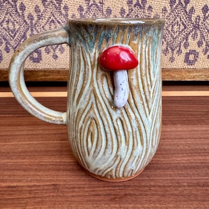 May include: Handmade ceramic mug with a textured, wood-grain design. The mug features a 3D red mushroom accent. The handle and rim have a brown and green glaze. A unique, nature-inspired coffee mug.