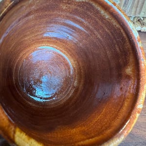 May include: A ceramic mug with a glossy, deep brown interior glaze that transitions to a lighter, mottled tan and cream finish along the thick, rounded rim. The surface shows visible wheel-thrown ridges and a smooth, reflective texture throughout.