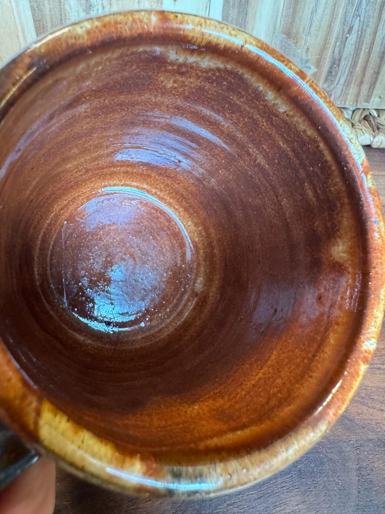 May include: A ceramic mug with a glossy, deep brown interior glaze that transitions to a lighter, mottled tan and cream finish along the thick, rounded rim. The surface shows visible wheel-thrown ridges and a smooth, reflective texture throughout.
