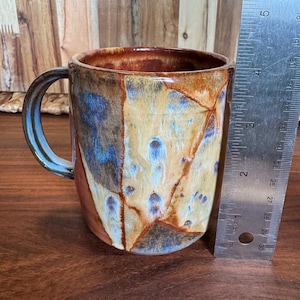 May include: A ceramic mug with a glazed finish in shades of tan, brown, and blue, featuring a curved handle. A metal ruler is positioned vertically next to the mug, indicating a height of approximately 10 cm from the base to the rim.