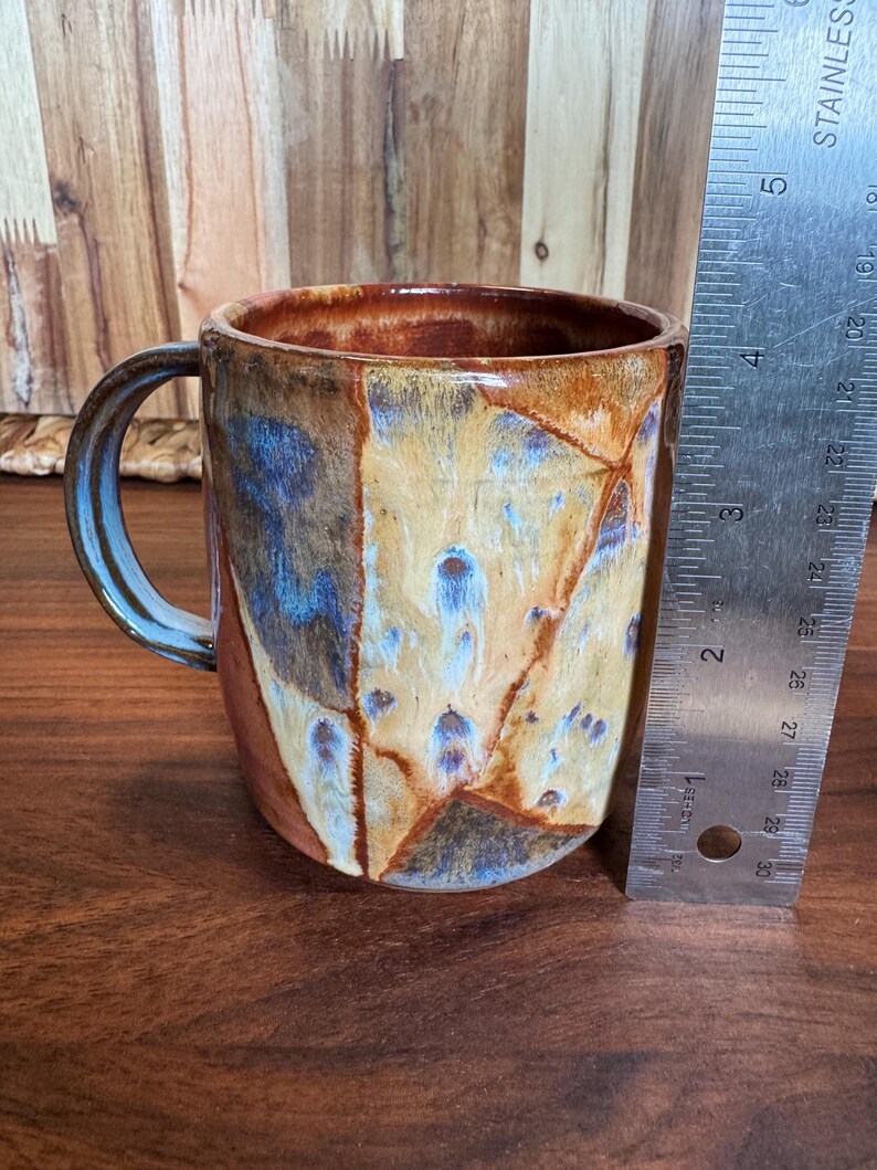 May include: A ceramic mug with a glazed finish in shades of tan, brown, and blue, featuring a curved handle. A metal ruler is positioned vertically next to the mug, indicating a height of approximately 10 cm from the base to the rim.