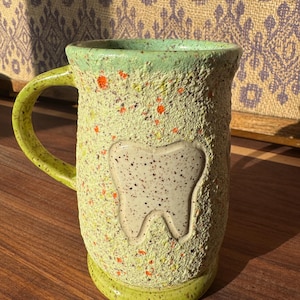 May include: A handmade ceramic mug with a textured, speckled green exterior and a light green handle and rim. A white tooth design is featured on the front. The mug is approximately 5 inches tall.