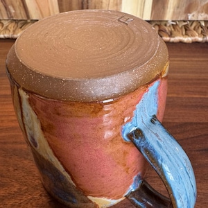 May include: A ceramic mug featuring a smooth, unglazed brown clay base at the top and a glossy, multi-coloured glaze on the body. The glaze displays swirls of pink, orange, and blue. A curved handle with blue and brown tones is attached to the side.