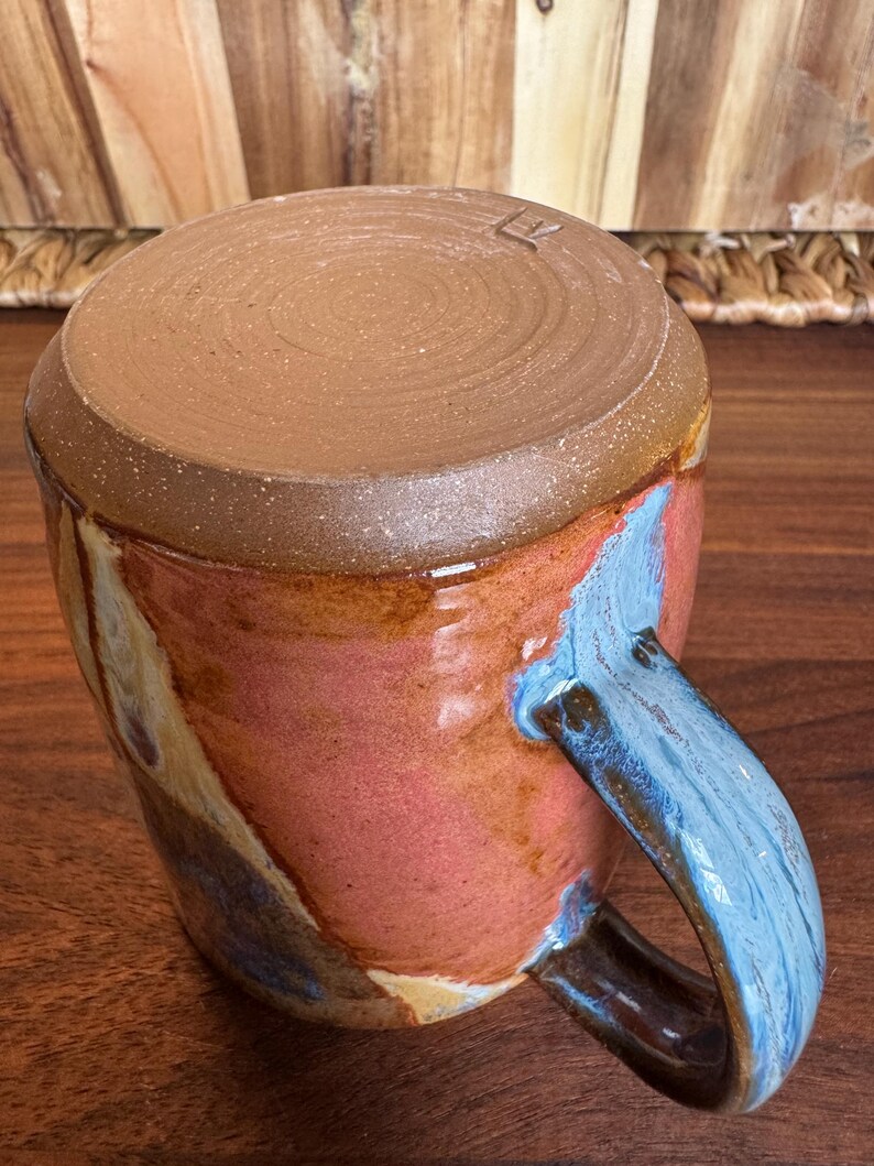 May include: A ceramic mug featuring a smooth, unglazed brown clay base at the top and a glossy, multi-coloured glaze on the body. The glaze displays swirls of pink, orange, and blue. A curved handle with blue and brown tones is attached to the side.