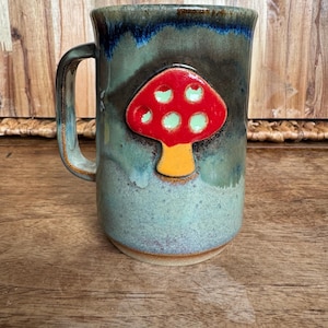 May include: A ceramic mug with a glossy glaze in shades of blue, green, and brown. The mug features a raised, three-dimensional applique of a mushroom with a bright red cap, light green spots, and a yellow stem, centered on the front surface.
