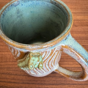 May include: Handmade ceramic mug with a unique design. The mug features a textured, ridged exterior with a brown and white color scheme. The interior is a teal-green color, and the handle is a matching brown. A small, green frog is attached to the side.