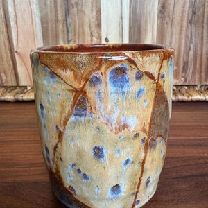 May include: A cylindrical ceramic tumbler featuring a glossy glaze with a mottled pattern of tan, brown, and deep blue hues. The surface displays a network of dark brown lines that create a segmented, crackled appearance across the exterior.
